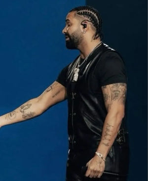 A man with braided hair, a beard, and visible arm tattoos is shown in profile against a solid blue background. He is wearing a black short-sleeve shirt and a black leather-like vest, with silver jewelry including a chain necklace and bracelet.