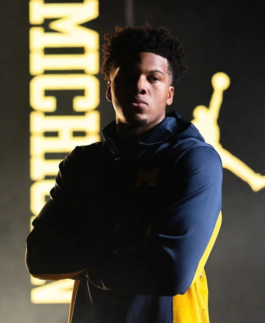 A man wearing a navy and yellow Michigan Wolverines hoodie stands with arms crossed, set against a dark background with a large yellow 'MICHIGAN' text and a Jordan logo.