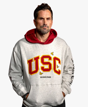 A man wearing a light gray heathered hoodie with a red hood lining and a large red and yellow 'USC' graphic on the front, with small butterfly accents and 'RYOKO RAIN' printed below.